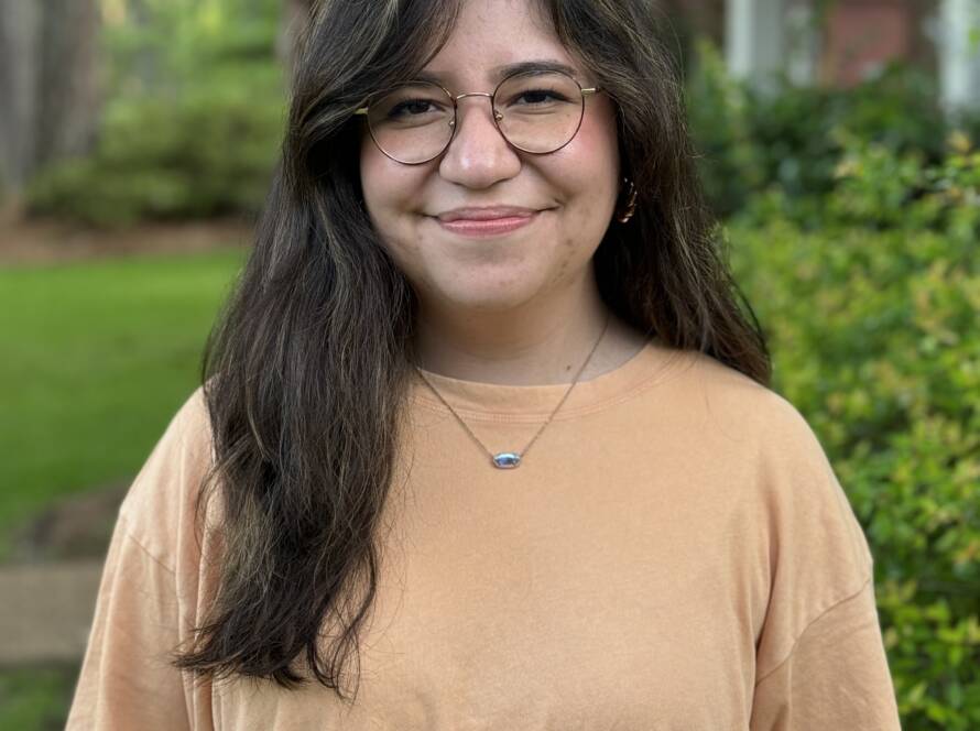 A woman with long hair and glasses standing in front of bushes.
